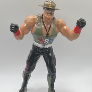 May include: A green, black, and red action figure of a wrestler wearing a green tank top with "USA" printed on the front, a black belt, black pants, and a green and black hat.