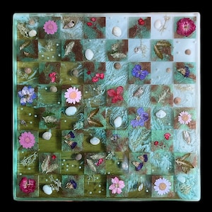 May include: A handcrafted chess board with a unique design. The squares are a mix of green, brown, and clear resin, adorned with dried flowers, seashells, and small pearls. The board is set against a black background.