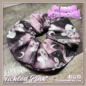 Puede incluir: Un scrunchie morado y negro con estampado de calavera y serpiente. El scrunchie está hecho de tela suave y tiene un diseño fruncido. El texto "Super Scrunchies" está impreso en la parte superior de la imagen. El texto "Tickled Pink" está impreso en la parte inferior de la imagen.