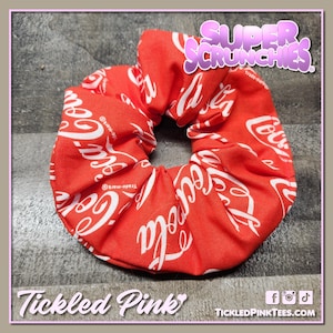 May include: Red scrunchie with a white Coca-Cola logo print. The scrunchie is a hair accessory, and the image includes the text "Super Scrunchies" and "Tickled Pink."