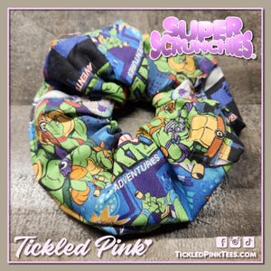May include: A fabric scrunchie featuring a colorful print of the Teenage Mutant Ninja Turtles. The scrunchie has a blue background with green, orange, and purple accents. The text "Adventures" is visible on the fabric. The scrunchie is from Tickled Pink.