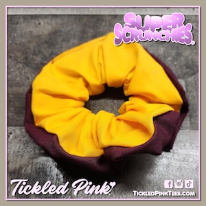 May include: A scrunchie with a gold center and a maroon border. The scrunchie is made of fabric and has the text "Super Scrunchies" in a pink, bubbly font. The text "Tickled Pink" is also visible.