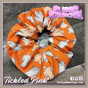 Op de afbeelding: Een oranje scrunchie van stof met een wit spookjespatroon. De scrunchie is gemaakt van een zachte, rekbare stof en heeft een label met de tekst "Super Scrunchies". De tekst "Tickled Pink" is ook zichtbaar op de afbeelding.