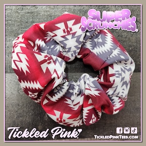 May include: A red, white, and gray patterned scrunchie with the text "Super Scrunchies" in purple letters. The scrunchie is made of fabric and is tied in a knot.