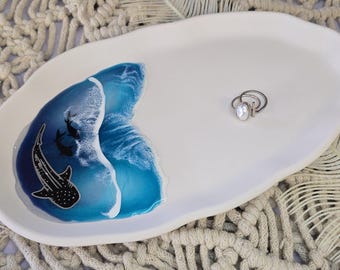 May include: White ceramic oval tray with a blue ocean wave design and a whale shark illustration. A silver ring with a white stone sits on the tray. The tray is on a macrame surface.