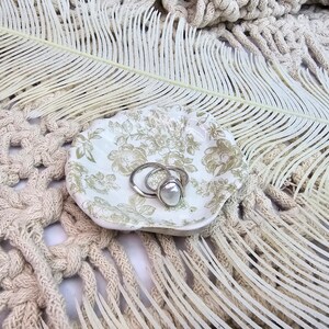 May include: A small, white, shell-shaped dish with a floral pattern holds two silver rings, one with a large, round, white pearl. The dish sits on a textured, cream-coloured surface with a feather and macrame.