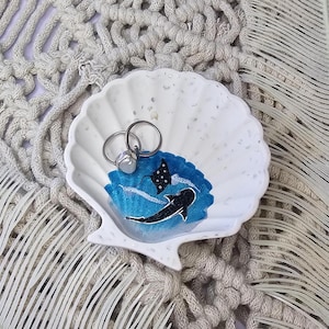 May include: White seashell-shaped trinket dish with a blue and white painted ocean scene featuring a manta ray and a shark. Two silver rings rest inside the dish. The dish is on a textured, off-white surface.