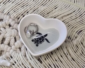 May include: A white, heart-shaped trinket dish featuring a black turtle design. A silver ring with a pearl bead rests inside. The dish is placed on a textured, off-white surface, with a feather-like decoration.