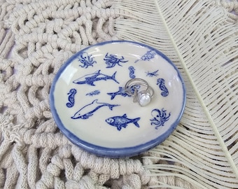 May include: A small, round ceramic dish with a blue rim and white interior. The dish is decorated with blue illustrations of various sea creatures, including sharks, fish, and seahorses. A silver ring with a white stone sits in the center. The dish is on a textured surface.