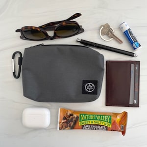 K&ouml;nnte beinhalten: Eine graue Rei&szlig;verschlusstasche mit einem kleinen Zahnradlogo, Sonnenbrille, ein Schl&uuml;sselbund mit Schl&uuml;sseln, Lippenbalsam, ein schwarzer Stift, eine braune Ledergeldb&ouml;rse, ein wei&szlig;es kabelloses Ladeetui und ein Nature Valley Sweet & Salty Nut dunkle Schokolade Erdnuss & Mandel M&uuml;sliriegel.