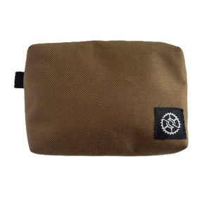 K&ouml;nnte beinhalten: Eine braune Stofftasche mit einem schwarzen Rei&szlig;verschluss und einem schwarzen Patch mit einem wei&szlig;en Zahnrad-Logo.