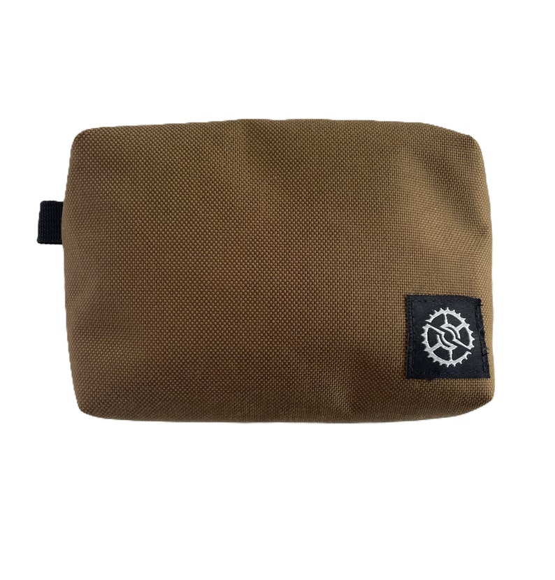 K&ouml;nnte beinhalten: Eine braune Stofftasche mit einem schwarzen Rei&szlig;verschluss und einem schwarzen Patch mit einem wei&szlig;en Zahnrad-Logo.