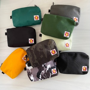 May include: Assortment of ten zippered pouches in various colors and patterns, including black, green, camouflage, and floral prints. Each pouch features a small flower design with a red center.