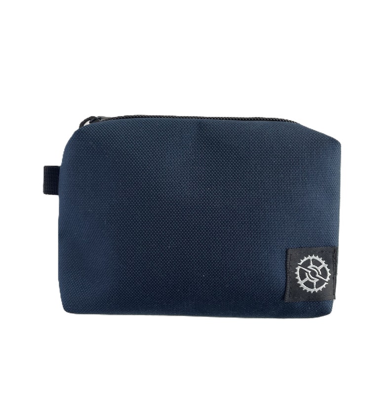 K&ouml;nnte beinhalten: Eine dunkelblaue Rei&szlig;verschlusstasche mit einem kleinen schwarzen Patch mit einem silbernen Zahnradlogo.