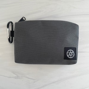 K&ouml;nnte beinhalten: Eine graue Rei&szlig;verschlusstasche mit einem kleinen schwarzen und wei&szlig;en Zahnradlogo. Die Tasche hat einen schwarzen Rei&szlig;verschluss und einen silbernen Karabinerclip.