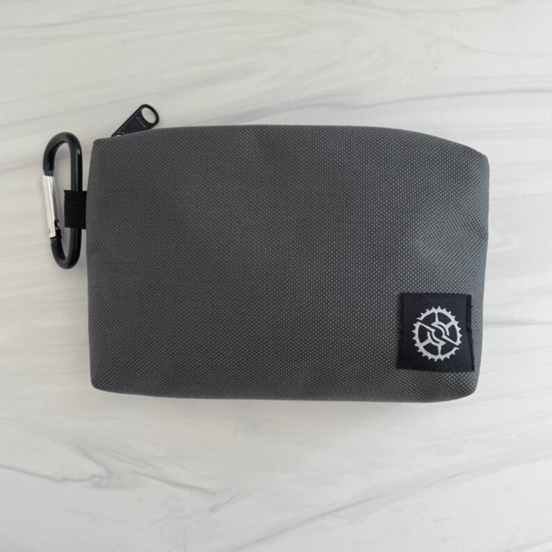 K&ouml;nnte beinhalten: Eine graue Rei&szlig;verschlusstasche mit einem kleinen schwarzen und wei&szlig;en Zahnradlogo. Die Tasche hat einen schwarzen Rei&szlig;verschluss und einen silbernen Karabinerclip.