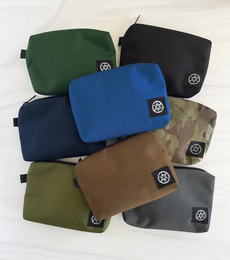 K&ouml;nnte beinhalten: Eine Sammlung von acht kleinen Rei&szlig;verschlusstaschen in verschiedenen Farben, darunter Gr&uuml;n, Blau, Braun, Schwarz und Tarnmuster. Jede Tasche hat ein kleines schwarz-wei&szlig;es Logo auf der Vorderseite.