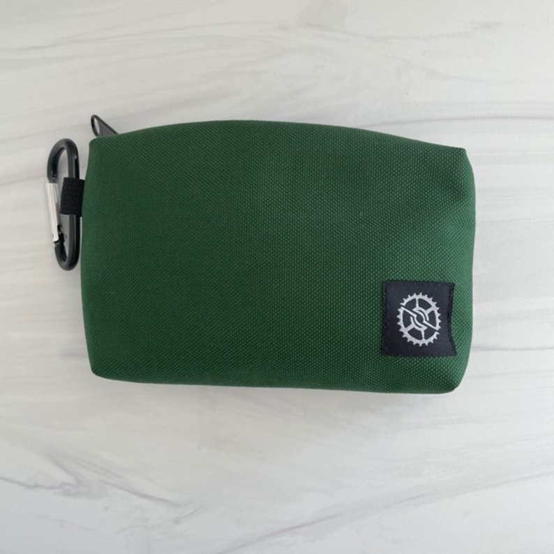 K&ouml;nnte beinhalten: Eine dunkelgr&uuml;ne Rei&szlig;verschlusstasche mit einem schwarzen Patch mit einem wei&szlig;en Zahnradlogo.