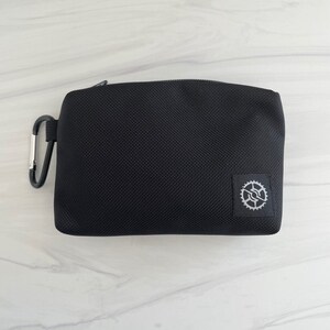 K&ouml;nnte beinhalten: Eine schwarze Rei&szlig;verschlusstasche mit einem kleinen silbernen Zahnradlogo. Die Tasche hat einen schwarzen Karabinerhaken.