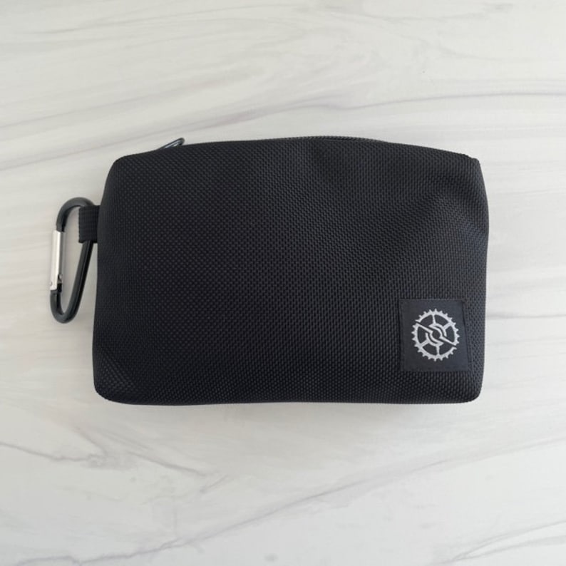 K&ouml;nnte beinhalten: Eine schwarze Rei&szlig;verschlusstasche mit einem kleinen silbernen Zahnradlogo. Die Tasche hat einen schwarzen Karabinerhaken.