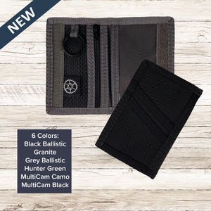 May include: A new, open wallet with a black exterior and a separate black card holder. The wallet has a zipper closure and a key ring. The text "NEW" is in the upper left corner. The text "6 Colors: Black Ballistic, Granite, Grey Ballistic, Hunter Green, MultiCam Camo, MultiCam Black" is in the lower left corner.