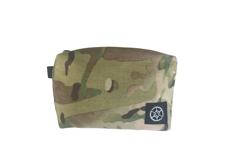 K&ouml;nnte beinhalten: Eine Tasche in Tarnmuster mit einem schwarzen Rei&szlig;verschluss und einem kleinen schwarzen Patch mit einem wei&szlig;en Logo in der unteren rechten Ecke.