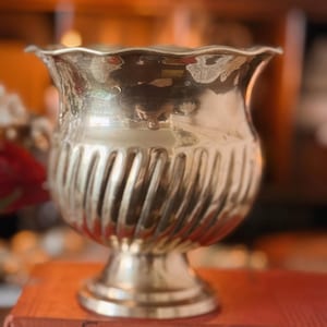 May include: A polished brass urn-shaped vessel with a scalloped rim and a spiraled, fluted body. The container features a pedestal base and sits atop a red book titled Old Curiosity Shop by Dickens, displaying a reflective metallic surface.
