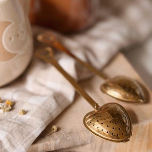 May include: Two gold-toned metal tea infusers with heart-shaped perforated heads and long, slender handles. Each handle features a small heart-shaped cutout at the end. The infusers have a polished, reflective surface with visible drainage holes.
