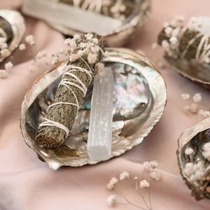 May include: Abalone shells with sage bundles and selenite crystals, arranged on a pink cloth. The sage bundles are wrapped with twine and adorned with small white flowers. The selenite crystals are rectangular and translucent.