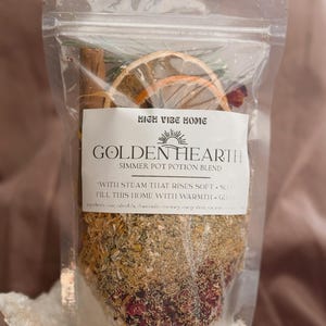 May include: A clear plastic bag containing a simmer pot potion blend. The label reads "HIGH VIBE HOME" and "GOLDEN HEARTH". The blend includes dried orange slices, cinnamon sticks, and various herbs and flowers. The text on the label says "WITH STEAM THAT RISES SOFT + SLOW FILL THIS HOME WITH WARMTH + GLOW".