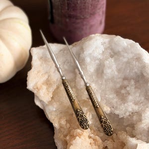 May include: Two metal picks with pointed silver tips and ornate, antique brass-coloured handles featuring embossed floral and leaf patterns. The handles have a textured, aged finish, while the slender shafts are smooth and metallic in appearance.