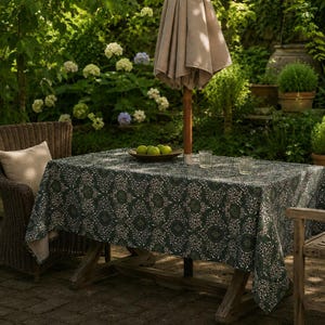 Puede incluir: Una mesa rectangular de exterior cubierta con un mantel verde oscuro con un estampado floral blanco. Un poste de madera para una sombrilla beige se encuentra en el centro, con un cuenco de madera con manzanas verdes y dos vasos transparentes.