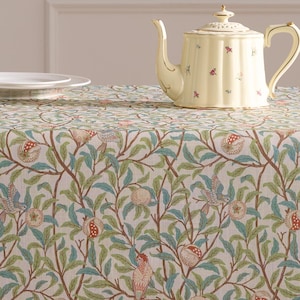 May include: A patterned tablecloth featuring a design of birds, leaves, and fruit in green, blue, and red on a cream backdrop. A yellow teapot and a white plate are on the cloth. The tablecloth covers a wooden table.