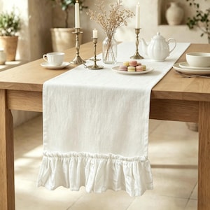 May include: A white linen table runner with a ruffled edge, displayed on a wooden table. The runner is adorned with a tea set, candles in brass holders, and a plate of colorful macarons, creating an elegant dining scene.
