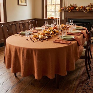 May include: An oval dining table covered with a solid burnt orange linen tablecloth. The table is set with clear glassware, green plates, and rust-colored cloth napkins. A centerpiece of dried autumn leaves, small pumpkins, and acorns runs down the center.
