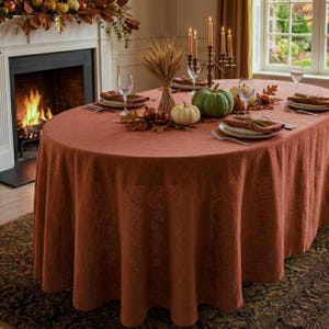 May include: An oval-shaped, solid rust-colored linen tablecloth draped over a dining table. The table is set with white plates, clear glassware, a metal candelabra with lit candles, a wheat sheaf centerpiece, and decorative pumpkins.