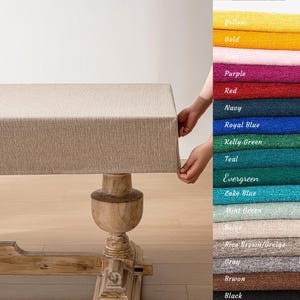 May include: A beige linen tablecloth draped over a wooden table with turned legs. The tablecloth has a subtle woven texture. A colour swatch card is shown on the right side of the image with various colours, including yellow, gold, purple, red, navy, royal blue, kelly green, teal, evergreen, lake blue, mint green, beige, rice brown/greige, grey, brown, and black.