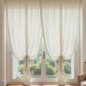 Op de afbeelding: Transparante, crèmekleurige gordijnen hangen voor een glazen deur. De gordijnen zijn in het midden vastgebonden, waardoor een gedrapeerd effect ontstaat. De deur onthult groen gebladerte buiten.