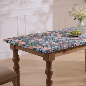 May include: A wooden dining table with a patterned tablecloth featuring a floral and animal design in shades of blue, green, and peach. The table has turned legs and is set with a vase of white flowers and a plate of green grapes.