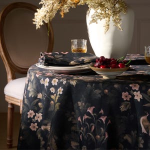 Puede incluir: Un mantel redondo con fondo oscuro y estampado floral en tonos rosa, beige y verde. Una servilleta y un plato a juego están sobre la mesa. También hay un jarrón blanco con flores secas y un cuenco con cerezas.
