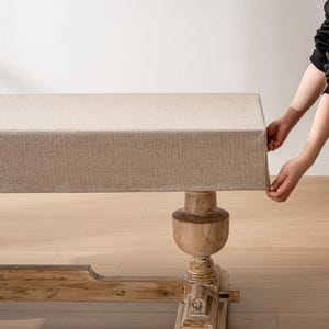 May include: A beige linen tablecloth is being placed on a wooden table with a carved pedestal base. The tablecloth has a subtle woven texture.