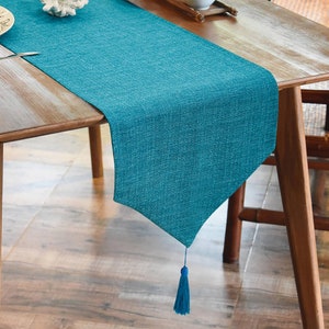 Puede incluir: Un camino de mesa de color turquesa con un borla en una mesa de madera. El camino de mesa tiene una textura de tejido tejido.