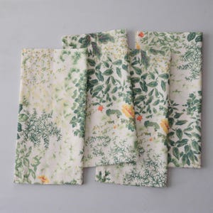 May include: Four white cloth napkins with a botanical print featuring green leaves, yellow flowers, and orange accents. The napkins are arranged in a slightly overlapping pattern, showcasing the detailed floral design.