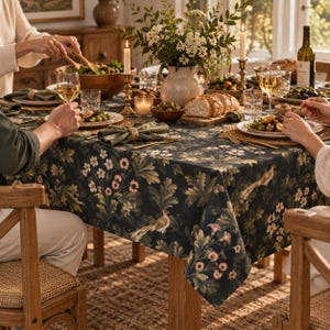 Dark Fern & Finch floral bird tablecloth draped over a wooden dining table, featuring a deep navy background with delicate botanical foliage, cream flowers, and vintage birds. Styled for an elegant autumn dinner with matching napkins, candlelight, and natural wood accents.