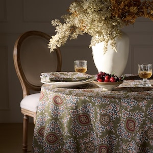 May include: A dining table set for a meal, featuring a round tablecloth with a green and red floral pattern. The table is set with white plates, green napkins, and clear glasses. A white vase holds dried flowers, and a bowl of cherries sits on the table.