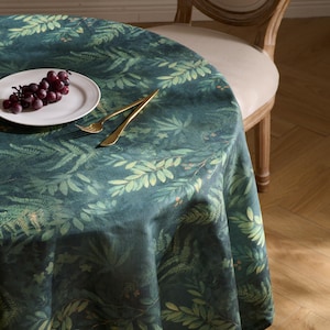 May include: A round tablecloth with a botanical print in shades of green and teal. The design features detailed illustrations of leaves and ferns. A white plate with grapes and gold cutlery are on the table. A wooden chair is in the background.