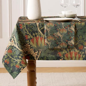 May include: A dark blue tablecloth with a floral pattern in shades of green, red, and beige. The tablecloth is draped over a wooden table, with a matching napkin and a white vase visible. The table is set with plates, bowls, and wine glasses.