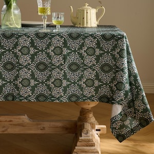 Pu&ograve; includere: Una tovaglia verde con un motivo floreale ripetuto copre un tavolo di legno. La tovaglia ha uno sfondo verde scuro con un motivo di disegni floreali bianchi, beige e verde scuro. Una teiera, bicchieri e un vaso di fiori sono sul tavolo.