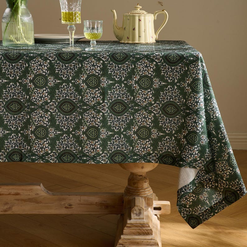Pu&ograve; includere: Una tovaglia verde con un motivo floreale ripetuto copre un tavolo di legno. La tovaglia ha uno sfondo verde scuro con un motivo di disegni floreali bianchi, beige e verde scuro. Una teiera, bicchieri e un vaso di fiori sono sul tavolo.