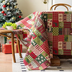 May include: A festive Christmas tablecloth featuring a patchwork design in red, green, and white. The pattern includes squares with reindeer, snowflakes, hearts, and polka dots. The tablecloth is draped over a wooden table, with a Christmas tree and gifts in the background.